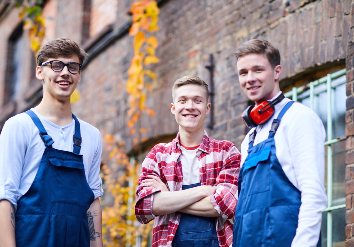 Drei junge sympathische Auszubildende im Blaumann vor einem Fabrikgebäude